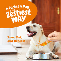 White dog sitting by metal bowl while person holds orange probiotic powder packet above it.
